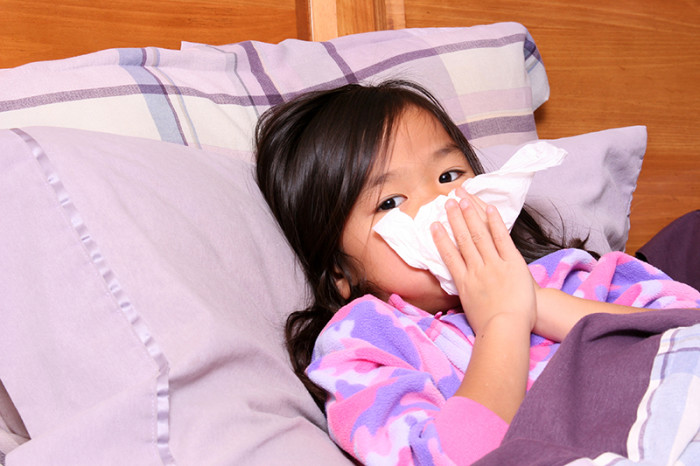Asian child lying in bed and holding tissue up to her runny nose