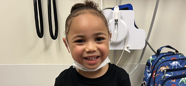 Lux, wearing a black shirt and a mask strapped under their chin, in a hospital setting.