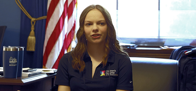 Riley, wearing a Children's Hospital of Richmond uniform in an office setting, american flag in the background.